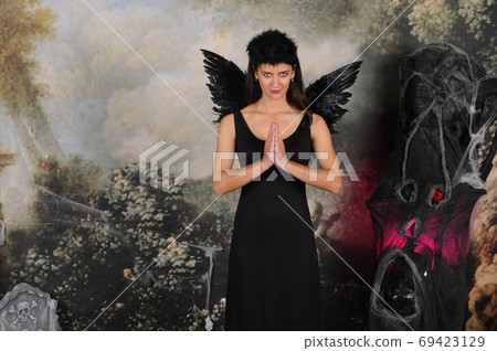 Beautiful young brunette woman in black angel costume with wings over dark spooky background in praying pose. Halloween party outfit 69423129