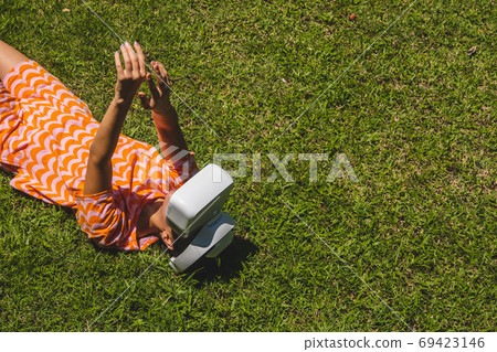 Young woman testing VR eyeglasses outside laying on grass. Female in bright orange summer outfit wearing virtual reality headset during summer. Active modern futuristic leisure lifestyle Young woman testing VR eyeglasses outside laying on grass. Female in bright orange summer outfit wearing virtual reality headset during summer. Active modern futuristic leisure lifestyle 69423146