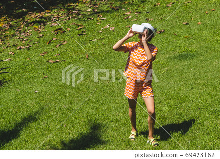 Young woman testing VR eyeglasses outside in tropical garden. Female in bright orange summer outfit wearing virtual reality headset during summer. Active modern futuristic leisure lifestyle Young woman testing VR eyeglasses outside in tropical garden. Female in bright orange summer outfit wearing virtual reality headset during summer. Active modern futuristic leisure lifestyle 69423162