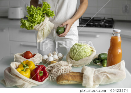 Woman's hand, holding a reusable grocery bag with vegetables on a kitchen at home and takes salad and avocado out. Zero waste and plastic free concept. Mesh cotton shopper with vegetables. Ecology. 69423374
