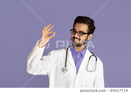 Smiling Indian doctor in lab coat holding pill on violet background Smiling Indian doctor in lab coat holding pill on violet background 69427519