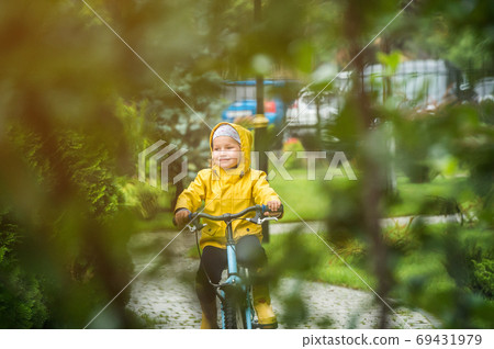 Happy little kid girl riding on bicycle in park. Child in waterproof yellow coat and rubber boots. Kid having fun on rainy day on bike. Playing on bike on sunny autumn rain day. 69431979
