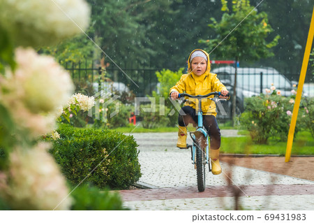 Cute little kid girl riding on bicycle in park. Child in waterproof yellow coat and rubber boots. Kid having fun on rainy day on bike. Playing on bike on sunny autumn rain day. Cute little kid girl riding on bicycle in park. Child in waterproof yellow coat and rubber boots. Kid having fun on rainy day on bike. Playing on bike on sunny autumn rain day. 69431983