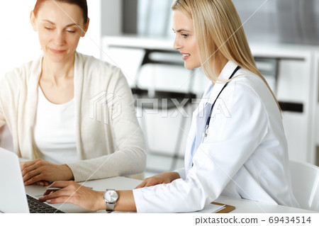 Woman-doctor at work in sunny hospital is happy to consult female patient. Blonde physician checks medical history record and exam results while using clipboard Woman-doctor at work in sunny hospital is happy to consult female patient. Blonde physician checks medical history record and exam results while using clipboard 69434514