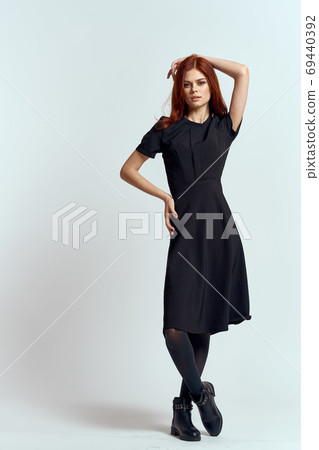 A woman in a black dress on a light background and pantyhose shoes red hair and pose in full growth 69440392