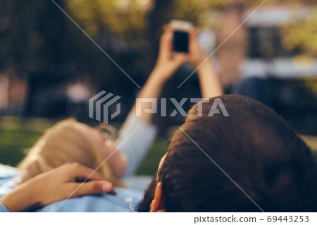 A man and a woman lie in the park on the grass with a phone in their hands rest communication A man and a woman lie in the park on the grass with a phone in their hands rest communication 69443253