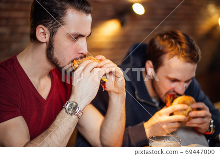 Two happy male friends eating tasty burgers in bar. Two happy male friends eating tasty burgers in bar. 69447000