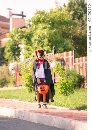 Little boy in costume of halloween devil with basket of treats standing on road 69450130
