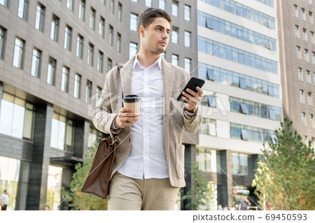 Busy young attractive manager with bag, drink and smartphone walking down street Busy young attractive manager with bag, drink and smartphone walking down street 69450593