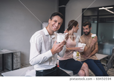 Dark-haired male holding chinese food box, his male and female colleagues sitting on table, having lunch Dark-haired male holding chinese food box, his male and female colleagues sitting on table, having lunch 69450594
