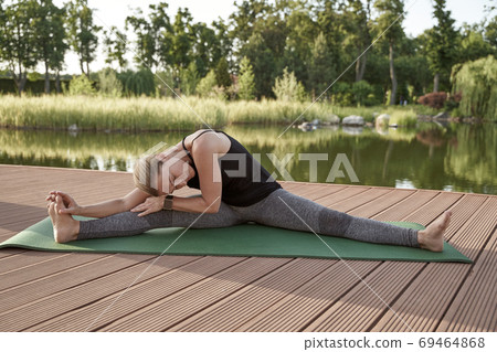 Stretching legs. Full length of a young beautiful fitness woman in sportswear doing twine exercise on yoga mat in park with small lake on a sunny summer morning 69464868