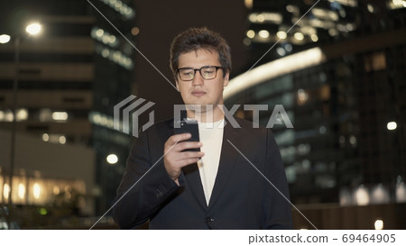 Businessman in eyeglasses scrolling the phone surrounded by buildings 69464905