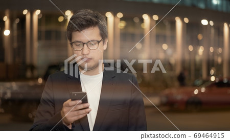 Businessman in eyeglasses scrolling the phone on background of building, lens flare 69464915