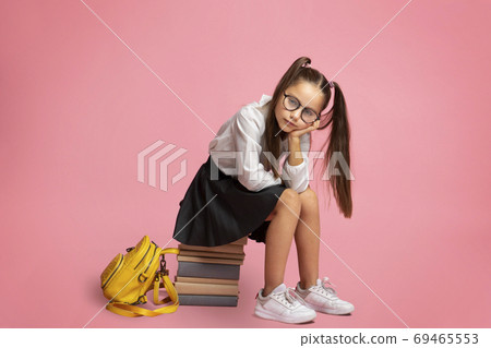 Problems in school education. Sad little schoolgirl in glasses with backpack sits on pile of books Problems in school education. Sad little schoolgirl in glasses with backpack sits on pile of books 69465553