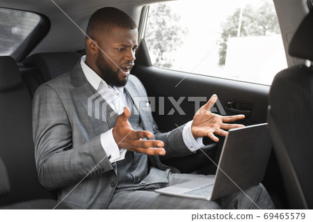 Angry black businessman with laptop going by taxi to job 69465579