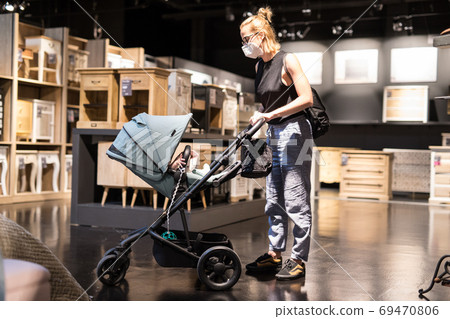Young mom with newborn in stroller shopping at retail furniture and home accessories store wearing protective medical face mask to prevent spreading of corona virus. New normal during covid epidemic Young mom with newborn in stroller shopping at retail furniture and home accessories store wearing protective medical face mask to prevent spreading of corona virus. New normal during covid epidemic 69470806