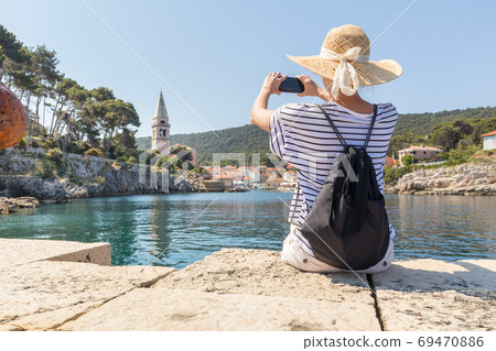 Woman traveler wearing straw summer hat and backpack, sittingat edge of stone pier, taking photo of beautiful panoramic view of Veli Losinj, Losinj island, Croatia 69470886