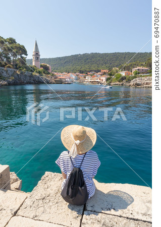 Woman traveler wearing straw summer hat and backpack, sittingat edge of stone pier, enjoying beautiful panoramic view of Veli Losinj, Losinj island, Croatia. Woman traveler wearing straw summer hat and backpack, sittingat edge of stone pier, enjoying beautiful panoramic view of Veli Losinj, Losinj island, Croatia. 69470887
