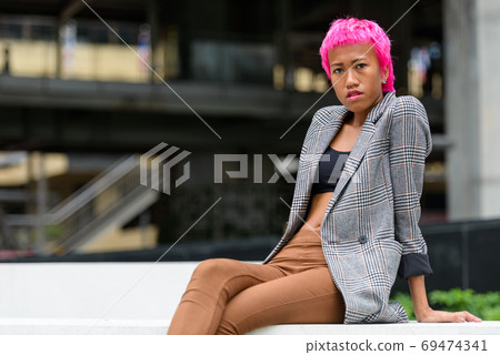 Young rebellious Asian businesswoman with pink hair sitting in the city Young rebellious Asian businesswoman with pink hair sitting in the city 69474341