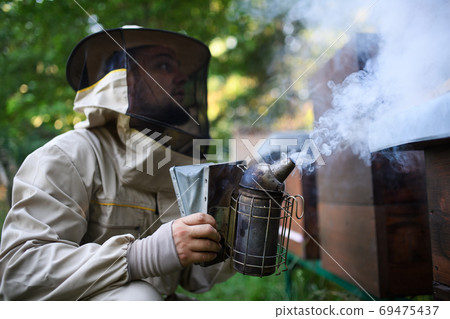 Portrait of man beekeeper working in apiary, using bee smoker. Portrait of man beekeeper working in apiary, using bee smoker. 69475437