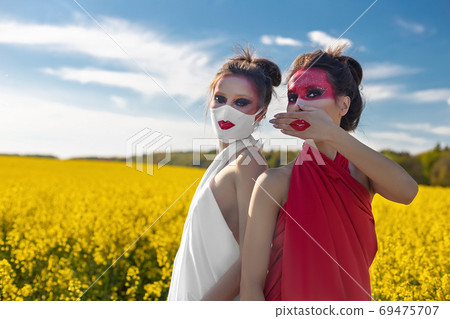 Two beautiful young brunette girls with creative bright makeup in tunics against a blue sky in a mask Two beautiful young brunette girls with creative bright makeup in tunics against a blue sky in a mask 69475707