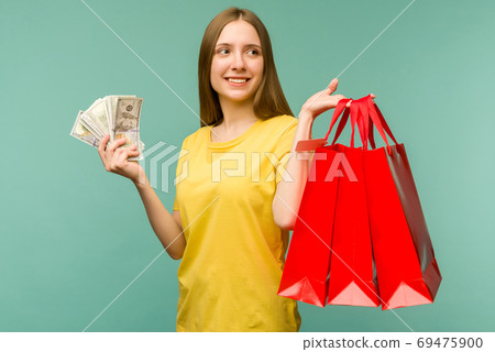 Photo of cheerful young woman holding fan of money and red shopping bags,isolated on blue background Photo of cheerful young woman holding fan of money and red shopping bags,isolated on blue background 69475900