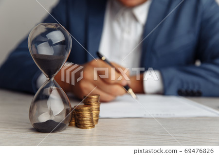 Businessman approves important contract in office. Stack of coins and hourglass on desk close-up. Time is money concept 69476826