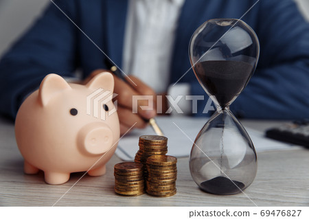 Businessman approves important contract in office. Pink piggy bank with stack of coins and hourglass on desk. Time is money 69476827