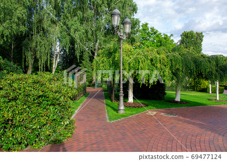 a well-kept park with pedestrian walkways paved with red tiles with many different plants, a lawn and trees with mulch, an iron lantern lighting on a sunny summer day. 69477124
