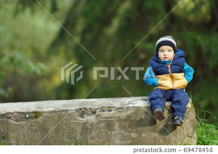 Thoughtful little boy in bright clothes sitting lonely on bench in park. Background of sunny green trees and bushes Thoughtful little boy in bright clothes sitting lonely on bench in park. Background of sunny green trees and bushes 69478458