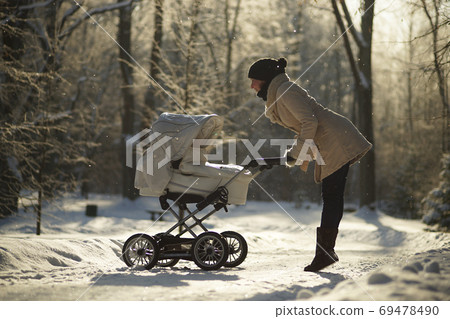 Young woman in coat raised on tiptoes, bending over baby stroller, looking at child. Sunny winter park, childcare, love Young woman in coat raised on tiptoes, bending over baby stroller, looking at child. Sunny winter park, childcare, love 69478490