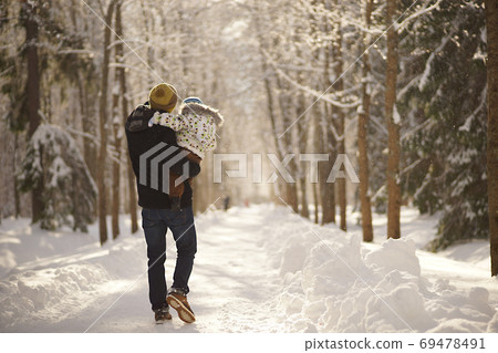 Young man with child on hands walking down the path in sunny winter park, snowy trees and snowbanks. View from behind Young man with child on hands walking down the path in sunny winter park, snowy trees and snowbanks. View from behind 69478491