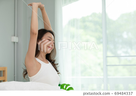 Woman stretching her arm after wake up on bed and looking outside windows in bedroom feeling so fresh and relax in the morning,Healthcare Concept 69484337