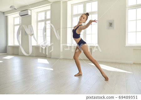 Young gymnast in black clothes with a ribbon performs an element of rhythmic gymnastics in the gym Young gymnast in black clothes with a ribbon performs an element of rhythmic gymnastics in the gym 69489351