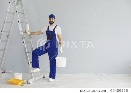 Smiling painter standing with paint roller and bucket in apartment for repairing and looking at camera 69489356