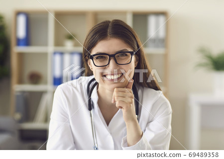 Portrait of friendly young female physician at desk in office smiling to patient 69489358