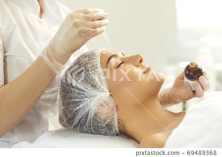 Professional cosmetician using pipette to apply moisturizing serum on client's face Professional cosmetician using pipette to apply moisturizing serum on client's face 69489368