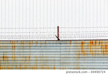 Barbed wire stretched over rusty brackets mounted on a fence made of metal sheets against a cloudy sky. 69490724