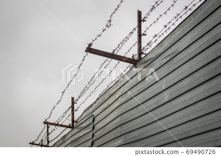 Barbed wire stretched over rusty brackets mounted on a fence made of metal sheets against a cloudy sky. 69490726