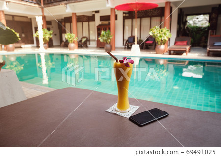 Mango smoothie with a smartphone on a sunbed against the background of the pool Mango smoothie with a smartphone on a sunbed against the background of the pool 69491025