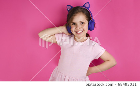 Beautiful little girl listening to music on color background. Pink clothes and a purple gadget. The Beautiful little girl listening to music on color background. Pink clothes and a purple gadget. The 69495011