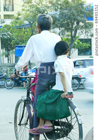 A Burmese man riding a bicycle with a girl sitting on the side of the road 69495352