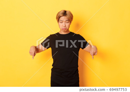 Portrait of gloomy and upset asian man sulking with regret, pointing and looking down at banner, yellow background 69497753