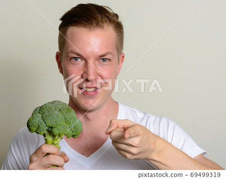 Portrait of happy young handsome man holding broccoli and pointing at camera Portrait of happy young handsome man holding broccoli and pointing at camera 69499319