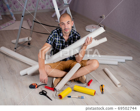 Portrait of a man on the floor with rolls of Wallpaper in his hands on the background of a stepladder. Portrait of a man on the floor with rolls of Wallpaper in his hands on the background of a stepladder. 69502744