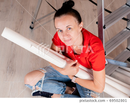 A happy woman with rolls of Wallpaper in her hands is sitting on the floor in the room in front of a stepladder. Renovation of the apartment. A happy woman with rolls of Wallpaper in her hands is sitting on the floor in the room in front of a stepladder. Renovation of the apartment. 69502801