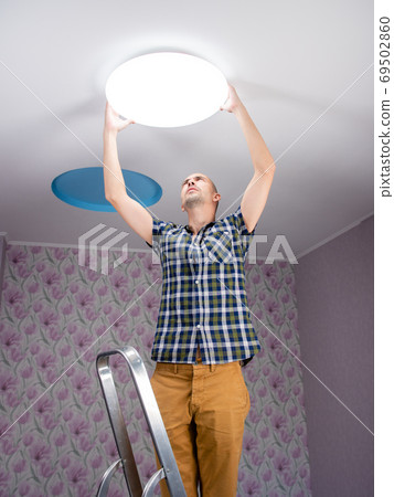 A man installs a modern led ceiling light on the ceiling. A man installs a modern led ceiling light on the ceiling. 69502860