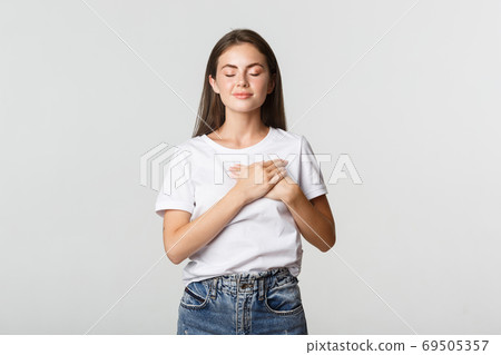 Portrait of tender and dreamy smiling woman, close eyes and holding hands on heart, daydreaming 69505357