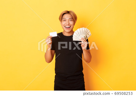 Young happy asian man with blond hair, showing credit card and money, smiling cheerful, standing yellow background Young happy asian man with blond hair, showing credit card and money, smiling cheerful, standing yellow background 69508253