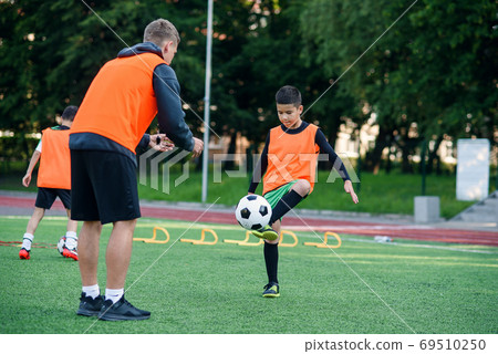 Motivated kid player in football uniform working out the kicking ball together with his experienced coach on sport field Motivated kid player in football uniform working out the kicking ball together with his experienced coach on sport field 69510250